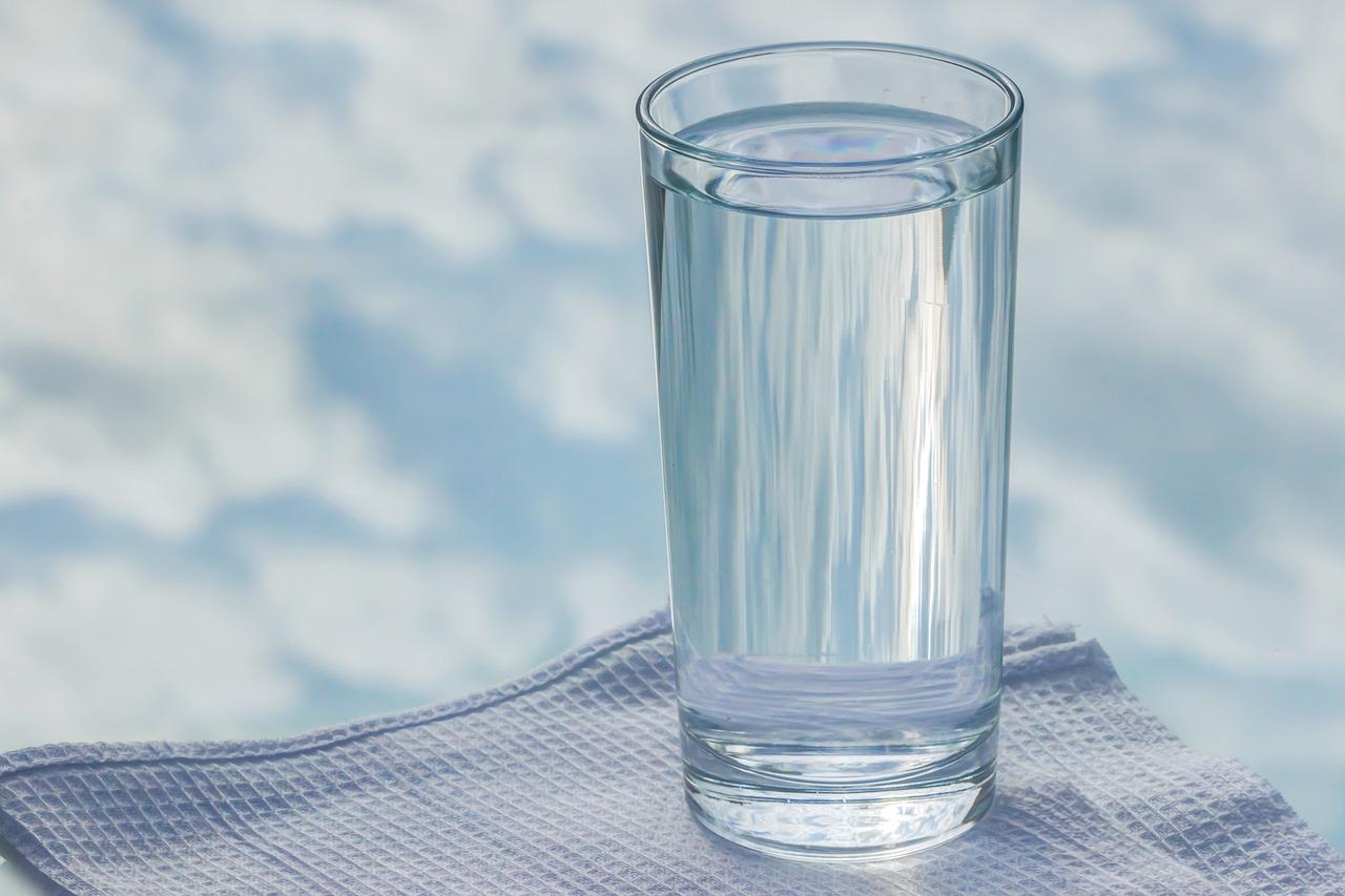 Foto de um copo de água. Um copo de vidro cheio de água está em cima de um guardanapo branco. O fundo da imagem também é branco.
